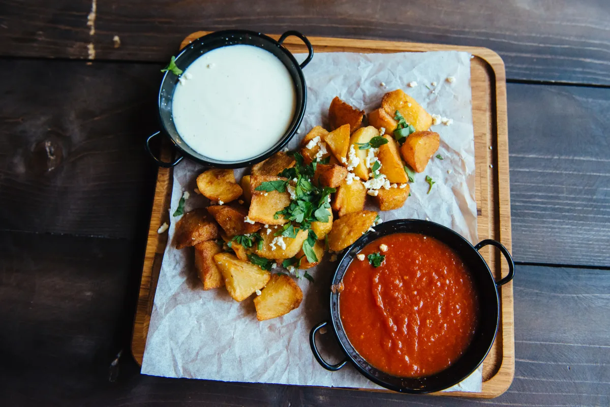 Frittierte Kartoffelwürfel mit Aioli und scharfer Tomatensauce auf einem Holzbrett