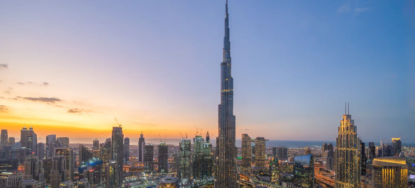 Der Burj Khalifa überragt die beleuchtete Skyline von Downtown Dubai, dahinter geht die Sonne über dem Meer unter