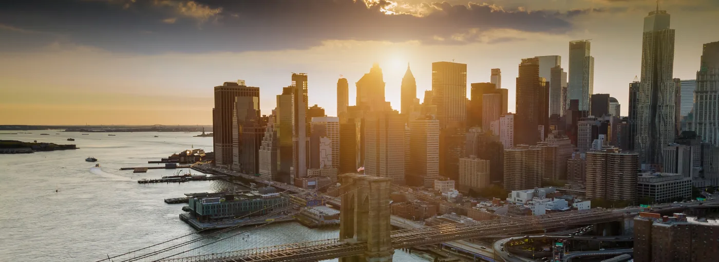Die Brooklyn Bridge im Abendlicht, dahinter die Hochhäuser von Lower Manhattan gegen die untergehende Sonne