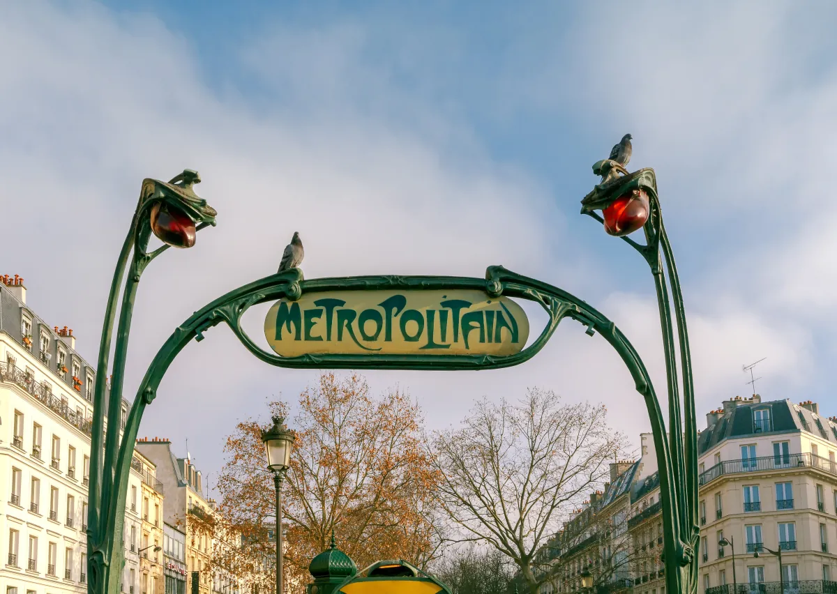 Ein Jugendstil-Eingang der Pariser Metro mit dem Schriftzug Métropolitain, dahinter Haussmann-Fassaden